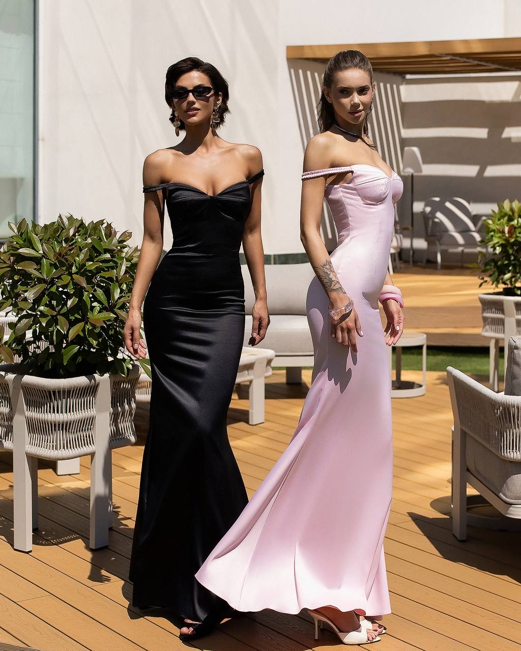 Two women in elegant evening gowns standing on a wooden deck with plants and chairs in the background.