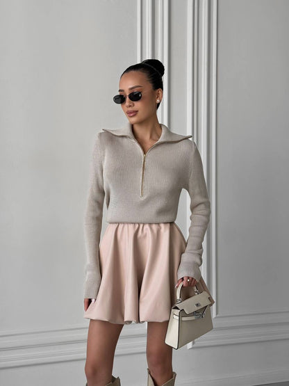 Woman wearing a beige sweater and pink skirt with sunglasses against a gray wall.