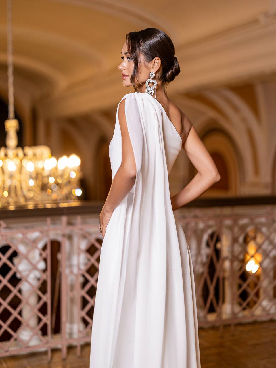 Woman in a white dress standing in an elegant indoor setting with chandeliers.