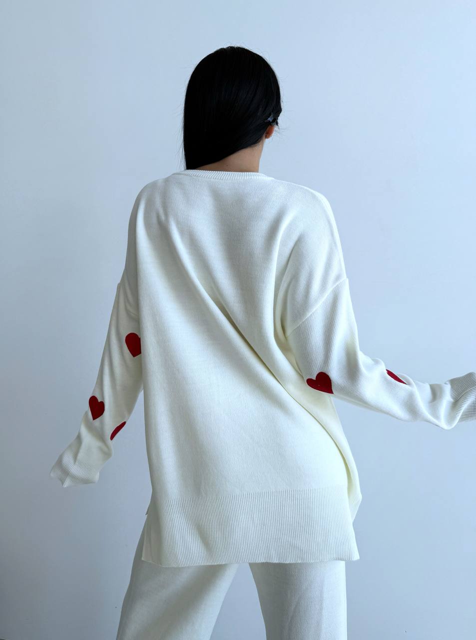 Person wearing a white sweater with red heart patterns on sleeves against a light gray background
