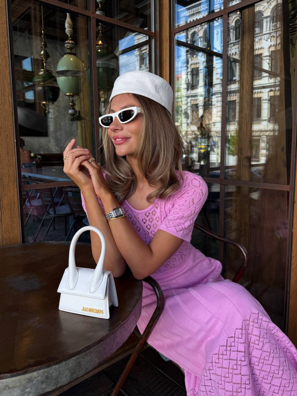 Woman in a pink dress sitting at a table with a white handbag, wearing sunglasses and a white cap.