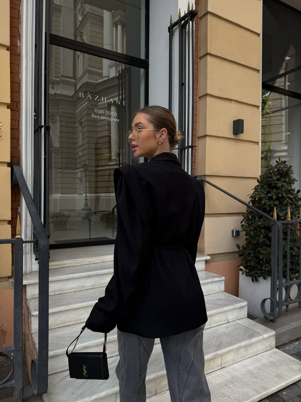Woman in black coat and gray pants standing on steps outside a building with 'Baztan' sign.