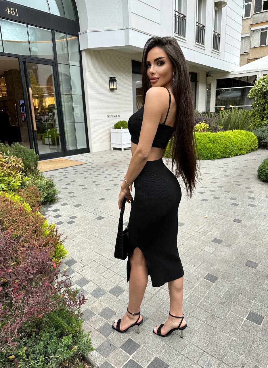 Woman in a black dress standing on a sidewalk with buildings and greenery in the background
