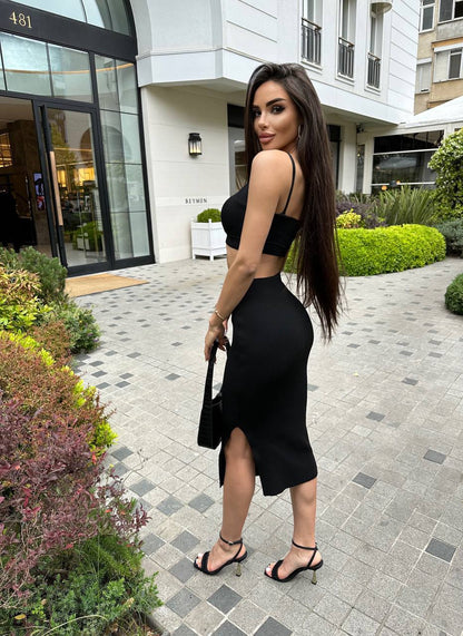 Woman in a black dress standing on a sidewalk with buildings and greenery in the background