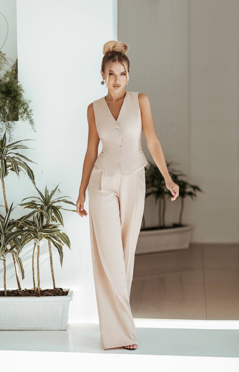 Woman in a beige sleeveless top and wide-leg pants standing indoors with plants in the background