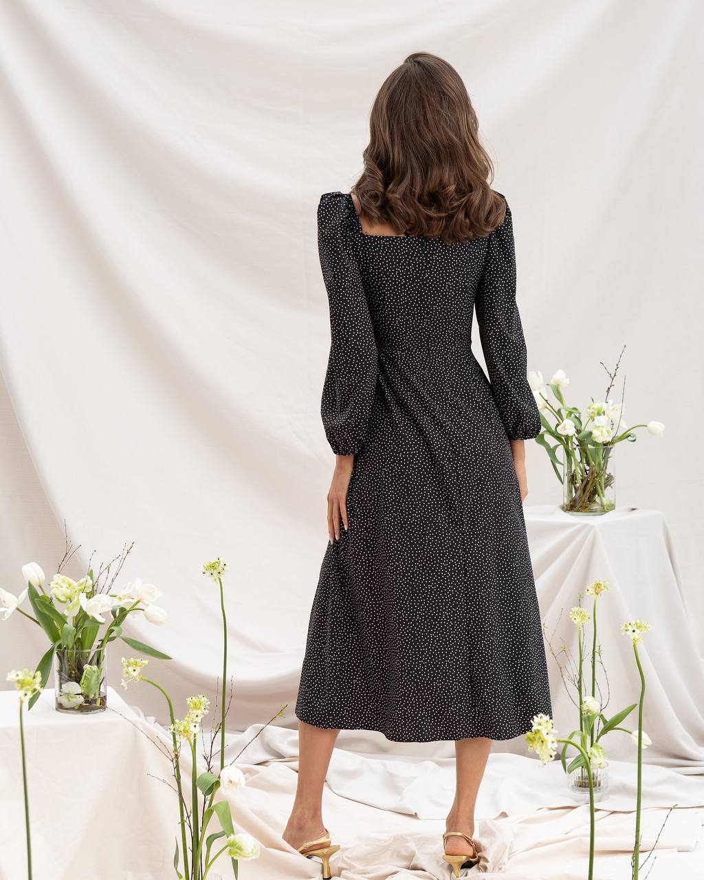 Woman wearing a black polka dot dress standing in front of a white curtain with floral decorations.