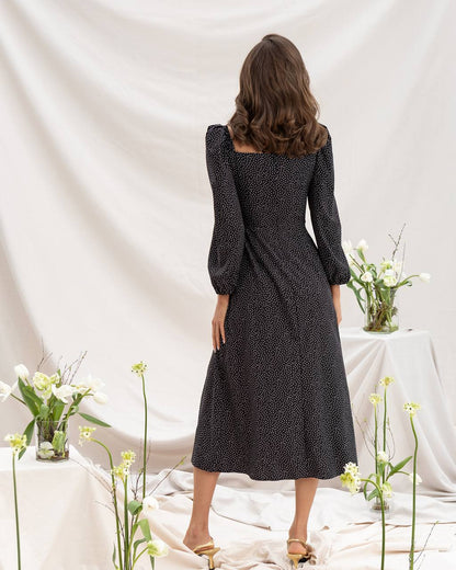 Woman wearing a black polka dot dress standing in front of a white curtain with floral decorations.
