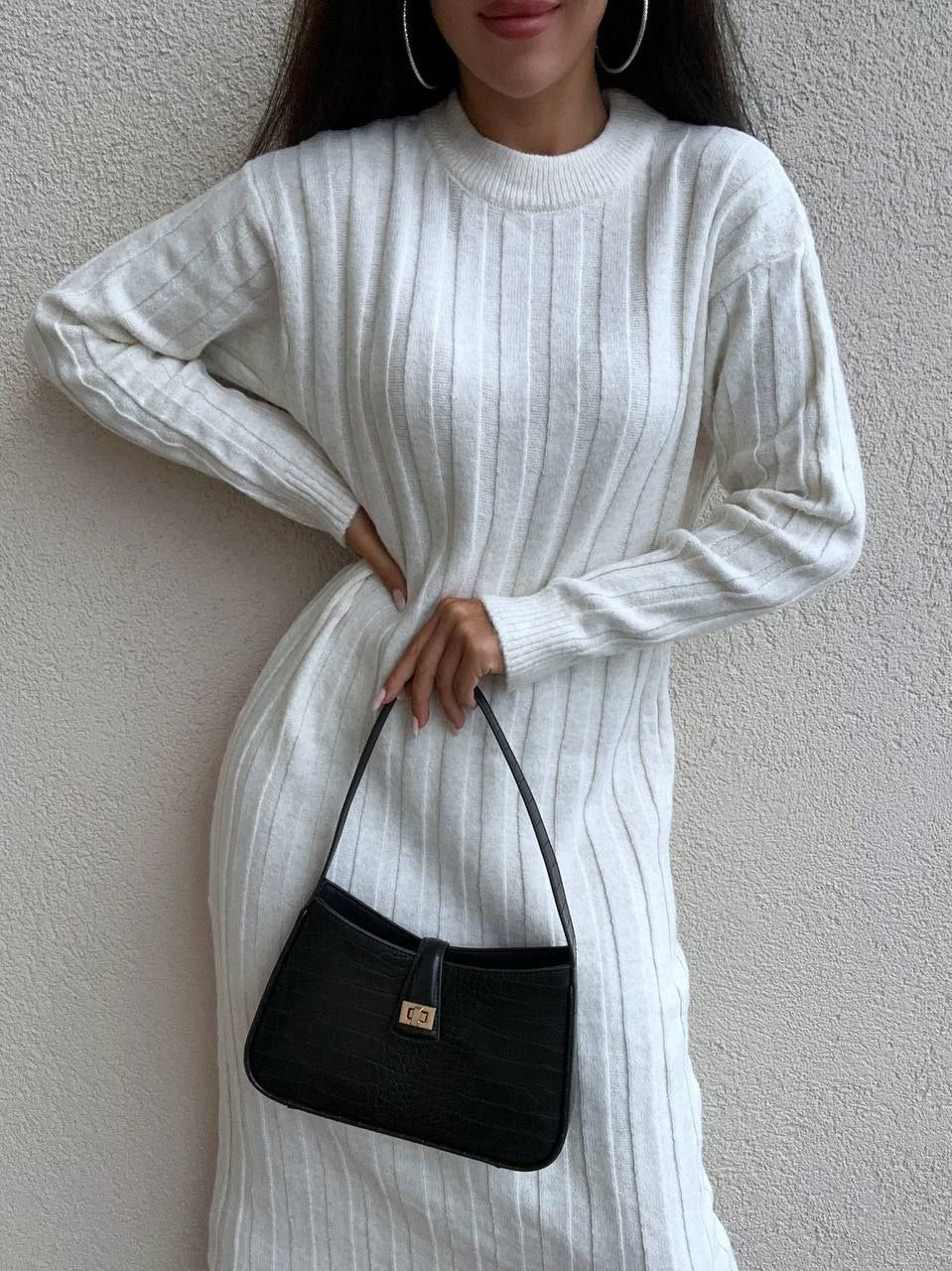 Person wearing a white ribbed sweater holding a black handbag against a plain background