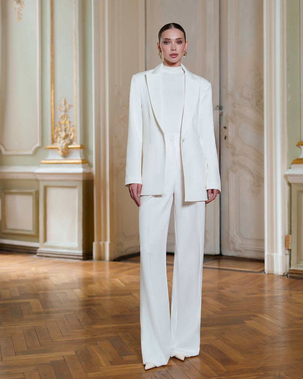 Person wearing a white suit standing in an elegant room with wooden flooring and decorative walls.