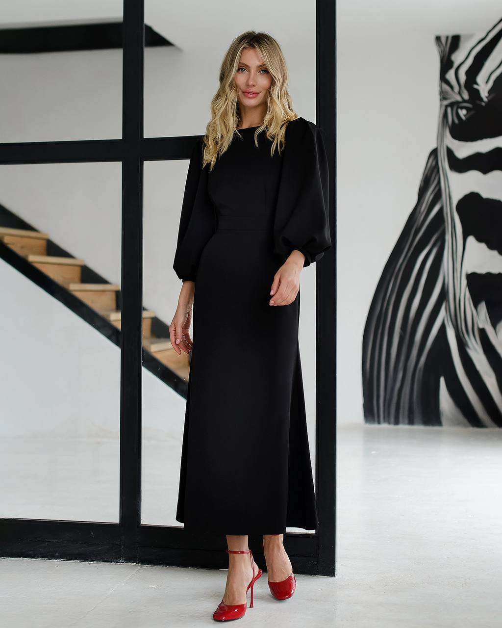 Woman in a black dress with red shoes standing in a modern interior setting.