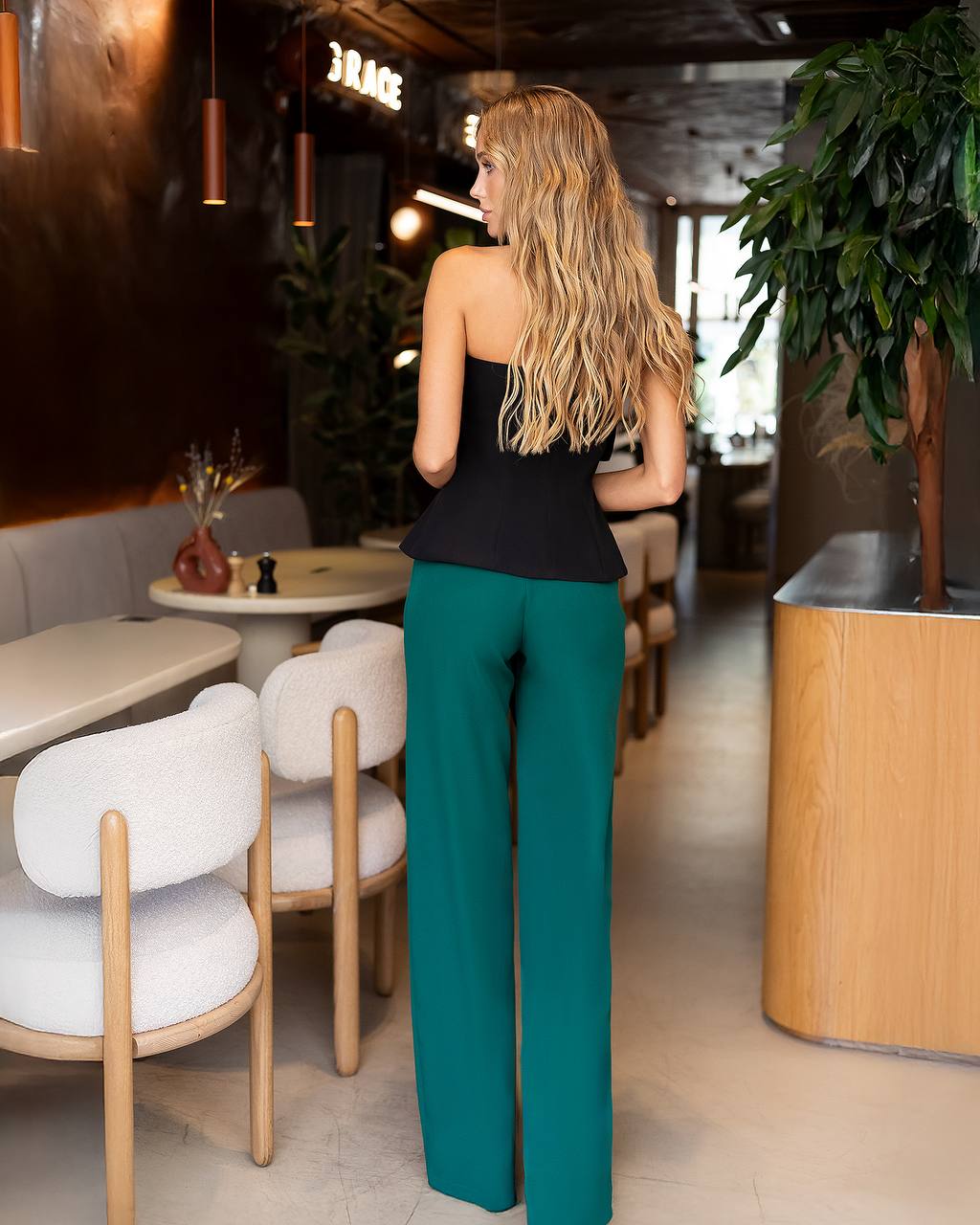 A woman with long blonde hair stands in a cafe, viewed from behind, wearing a black strapless peplum top and teal wide-leg trousers.