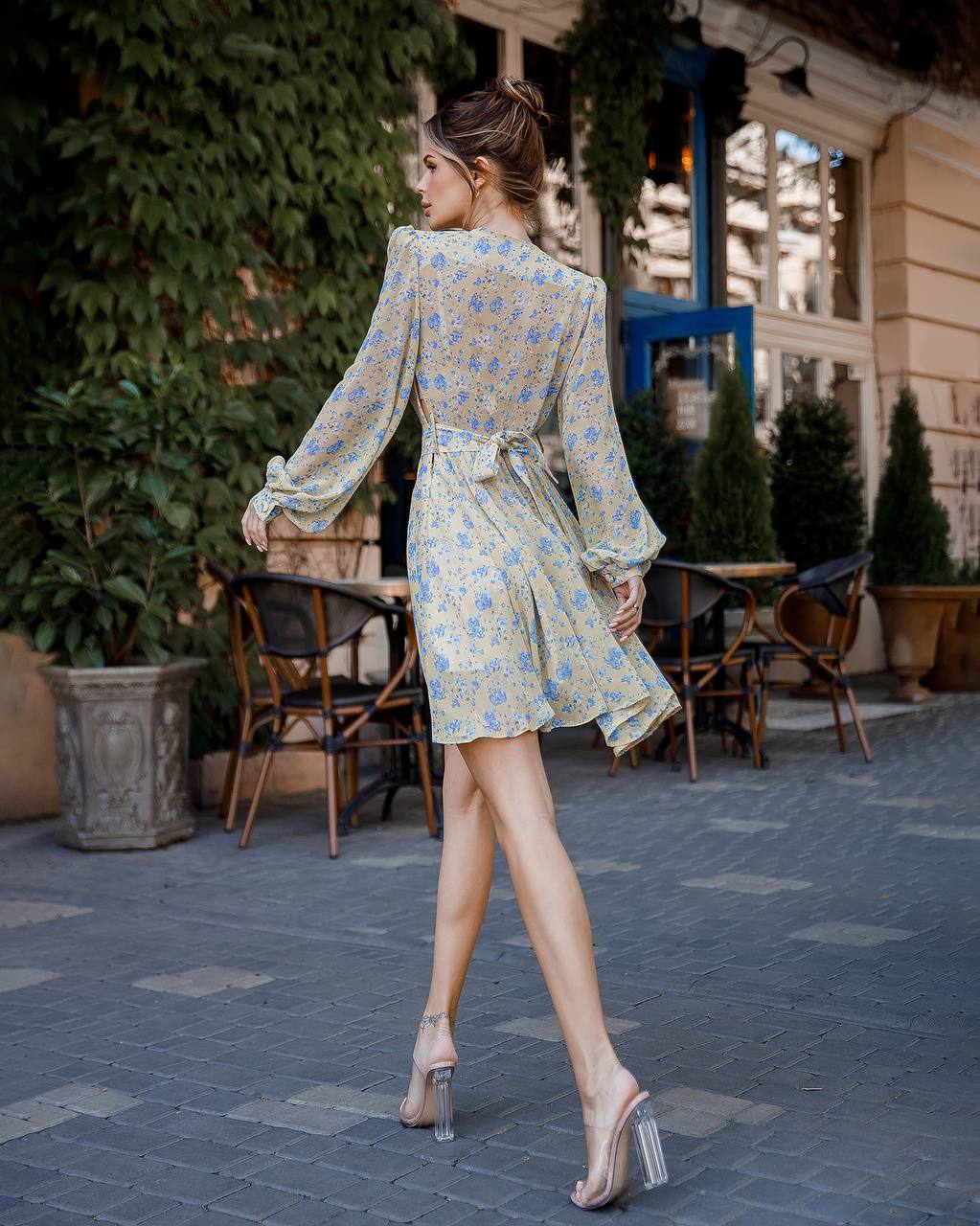 Woman in a floral dress walking outdoors on a street with plants and tables.