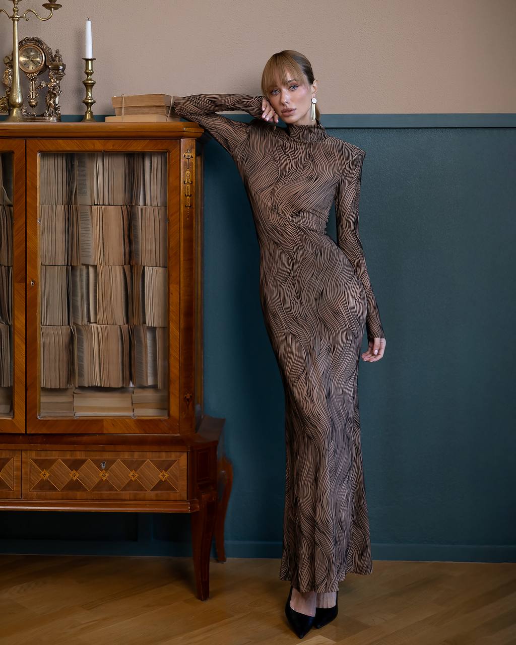 Woman in a patterned dress leaning against a wooden cabinet with a blue wall in the background
