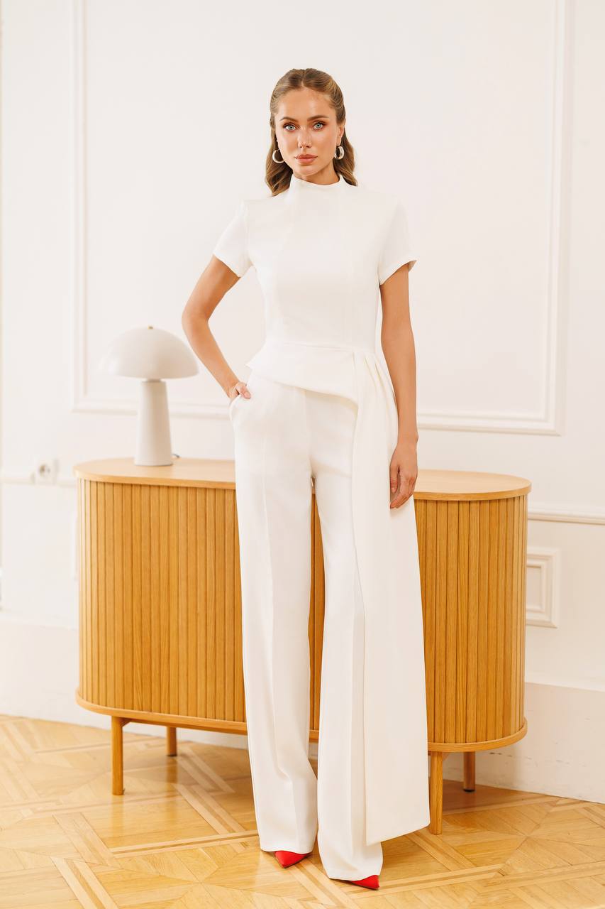 Woman in a white outfit standing in a room with wooden furniture and a white wall.