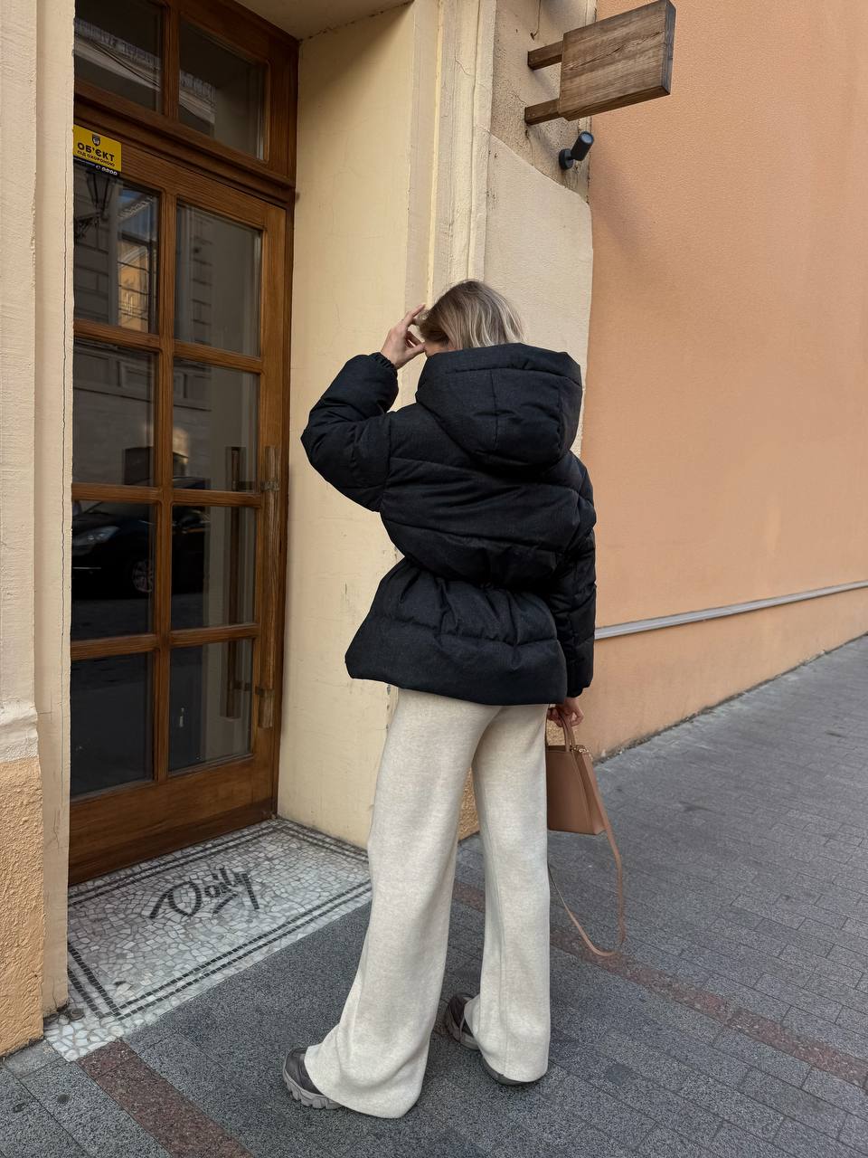 Person wearing a black puffer jacket and white pants standing on a street.