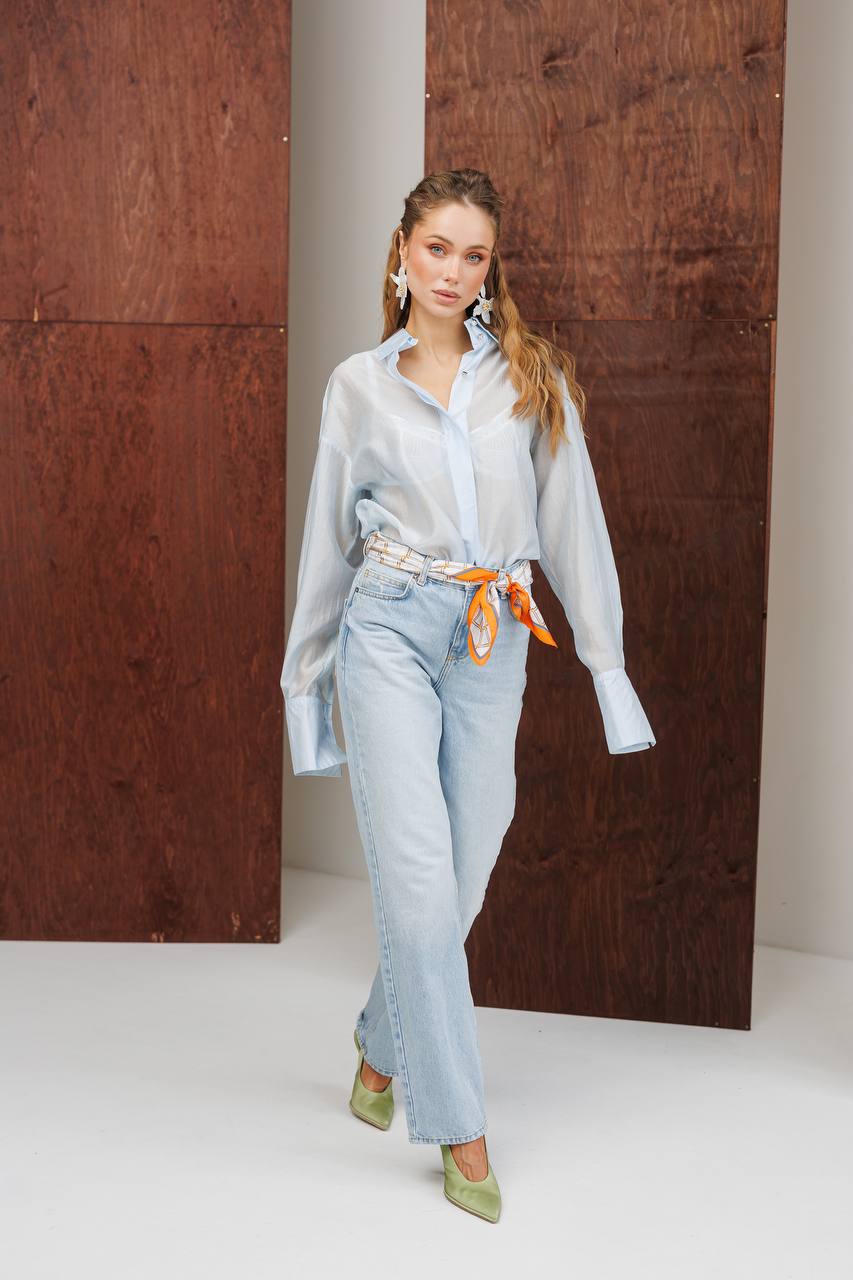 Woman wearing a light blue shirt and jeans with an orange belt, standing against a wooden panel background.