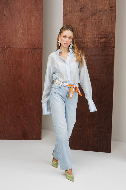 Woman wearing a light blue shirt and jeans with an orange belt, standing against a wooden panel background.