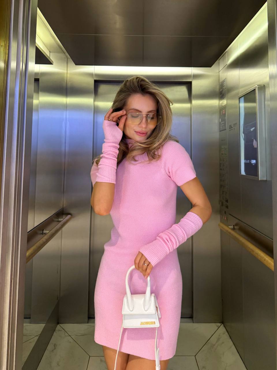 Woman in a pink outfit with matching gloves and a white handbag in an elevator.