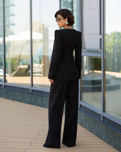 A woman stands on a modern outdoor terrace wearing a black tailored pantsuit with a structured blazer and wide-leg trousers, photographed from the back against large glass windows.