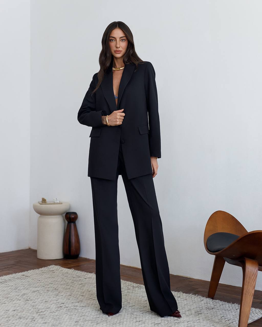 Woman wearing a black suit standing in a minimalistic room with a chair and decorative items.