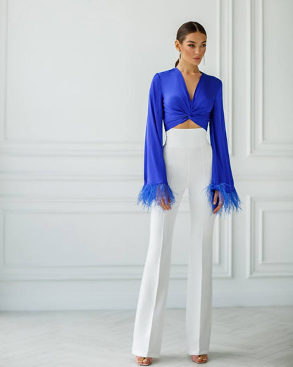 Woman wearing a blue top with fringed cuffs and white pants against a white wall.
