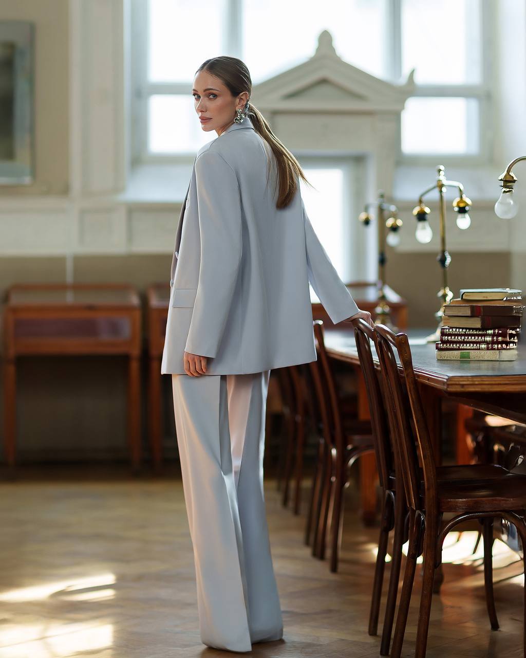 Woman in a light gray suit standing in a room with wooden furniture and large windows.