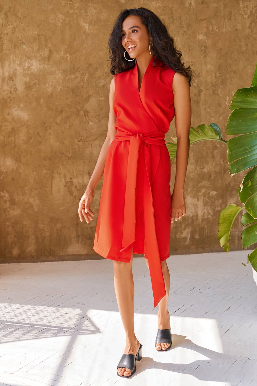 Woman wearing a red dress standing against a textured wall with a plant to the side.
