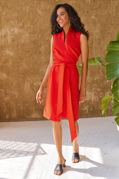 Woman wearing a red dress standing against a textured wall with a plant to the side.