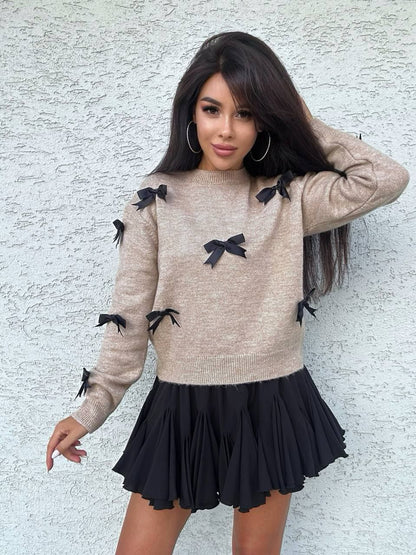 Woman wearing a beige sweater with black bow details and a black skirt against a light gray wall.