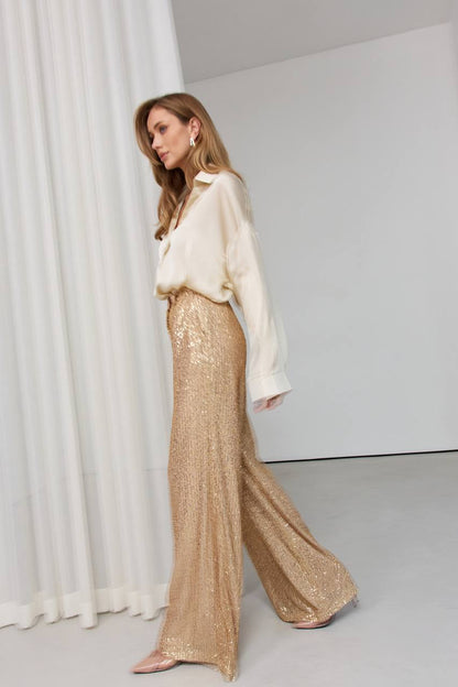 Woman wearing a white blouse and gold sequin pants against a white background