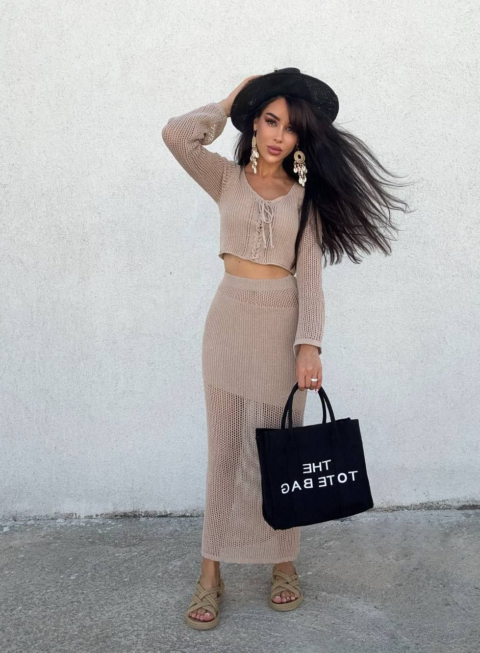 Woman in a beige outfit holding a black tote bag against a white wall