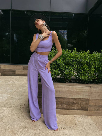 Woman in a lavender outfit posing outdoors with greenery and a building in the background
