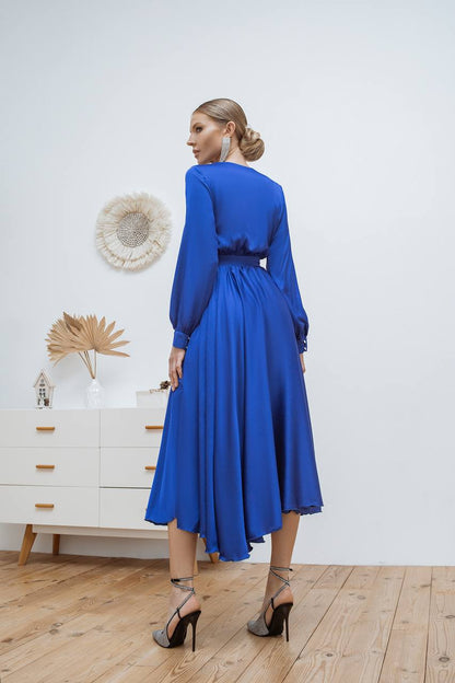 Woman in a blue dress standing in a room with a white wall and wooden floor.