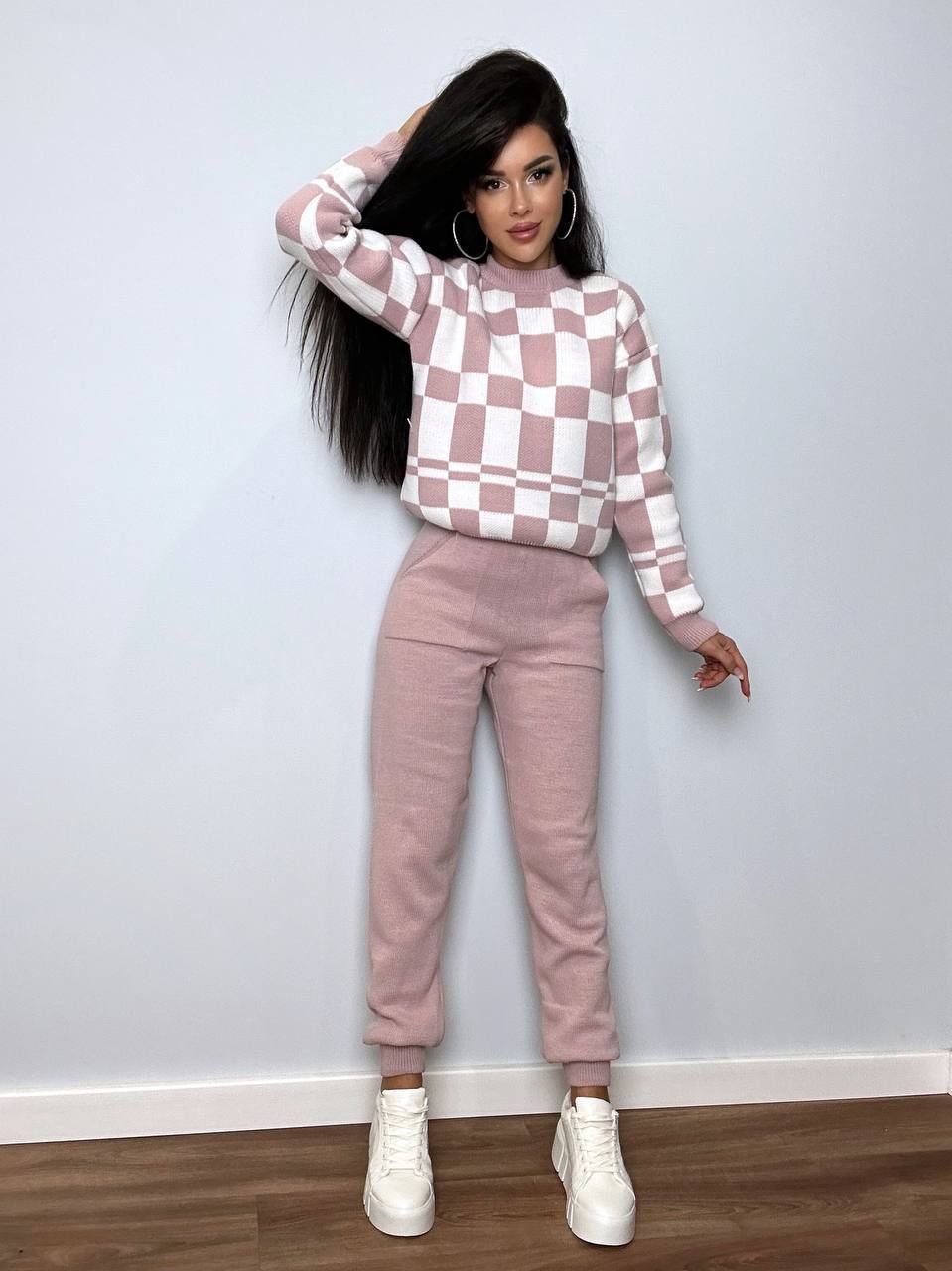 Woman wearing a checkered sweater and pink pants against a plain wall.