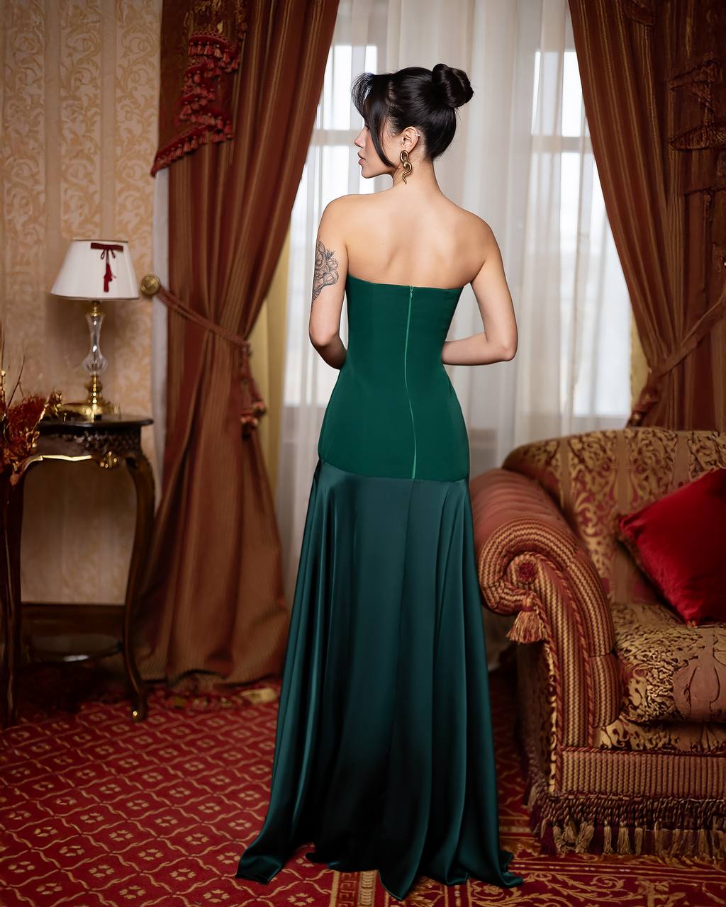 Woman in a green strapless gown standing in an elegant room with red carpet and patterned walls.
