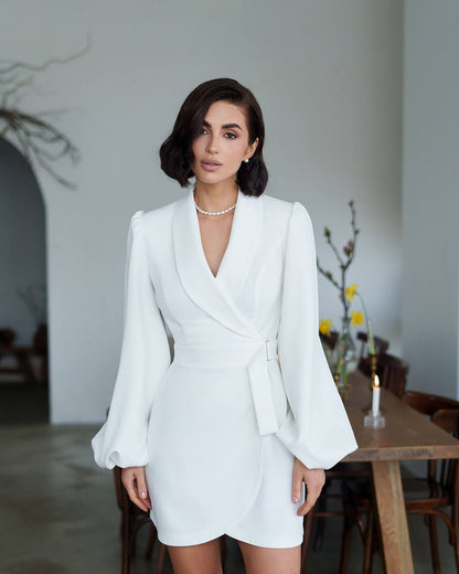 Woman in a white blazer dress standing in a room with a wooden table and decorative elements.
