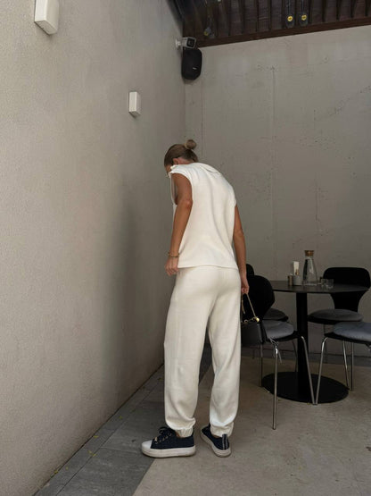 Person in a white outfit standing in a minimalistic room with tables and chairs.