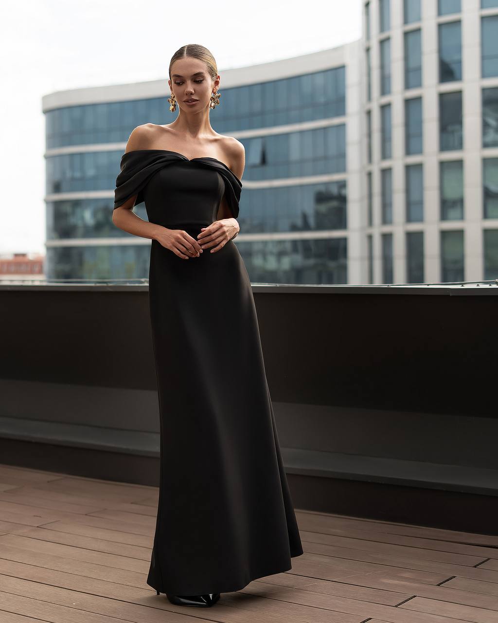 Woman in a black off-shoulder gown standing on a rooftop with modern building in the background
