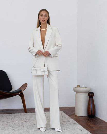 Woman wearing a white suit standing in a minimalistic room with a chair and table.