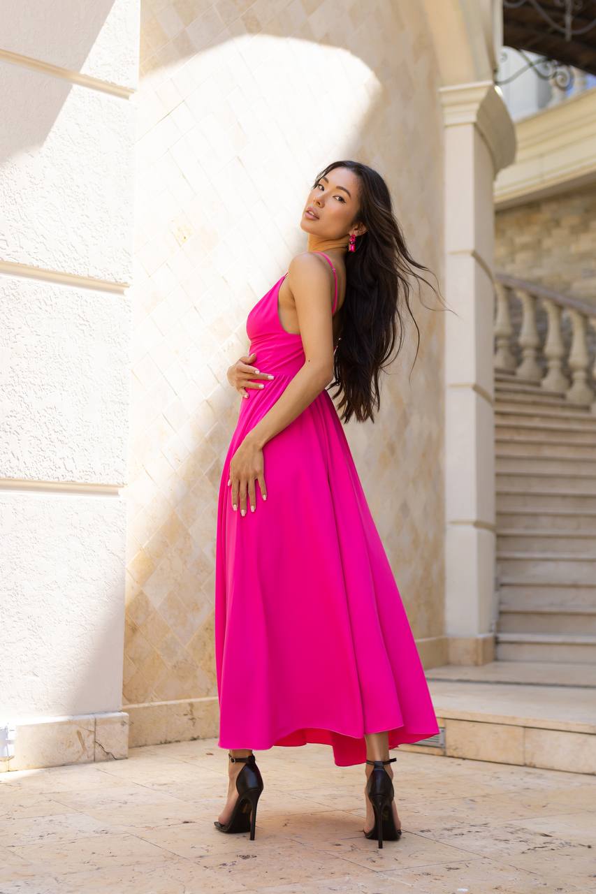 A woman posing in a vibrant hot pink halter-neck midi dress with black high-heeled boots, standing in front of a beige stone building with columns and stairs.