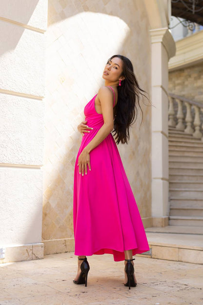 A woman posing in a vibrant hot pink halter-neck midi dress with black high-heeled boots, standing in front of a beige stone building with columns and stairs.