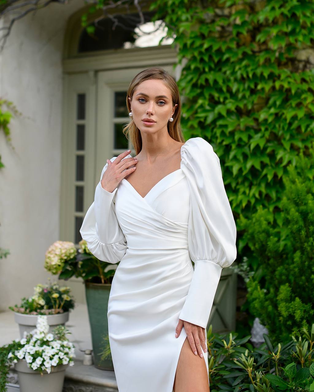 Woman in a white dress standing outdoors with greenery and a building in the background
