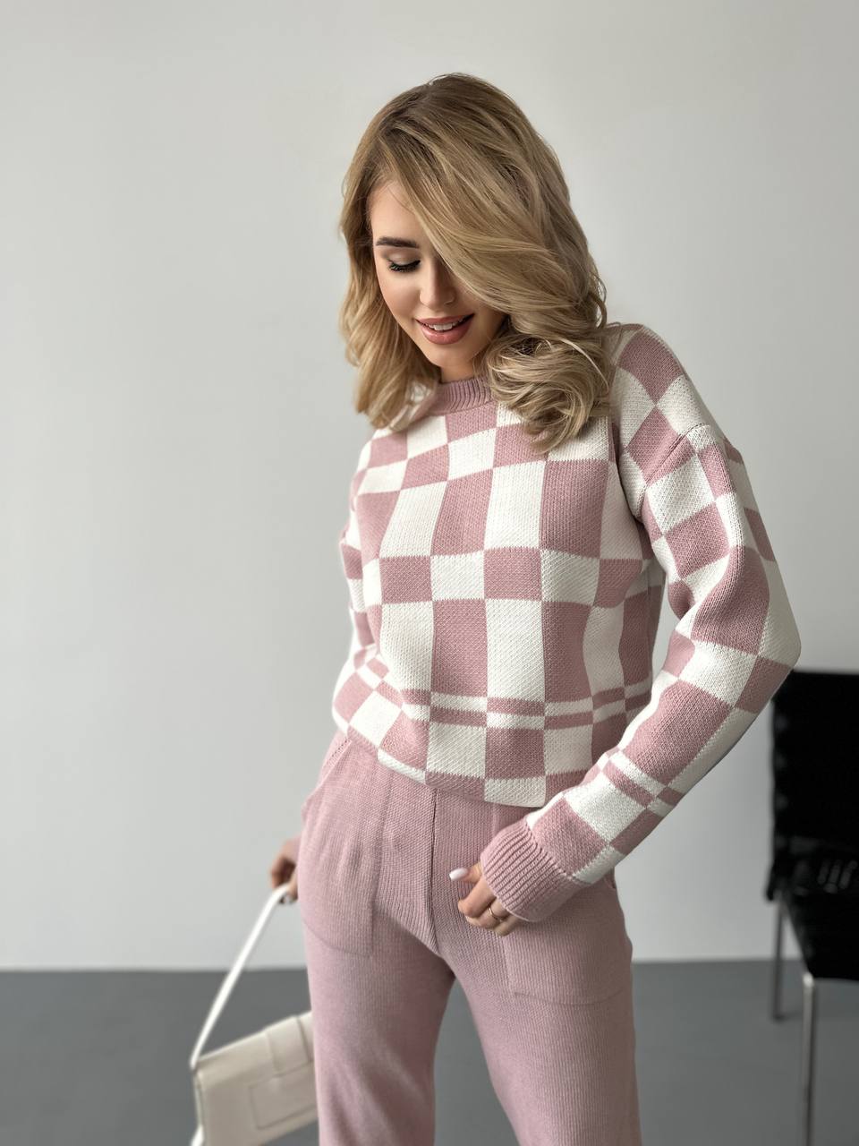 Woman wearing a pink and white checkered sweater and pants, holding a beige handbag.