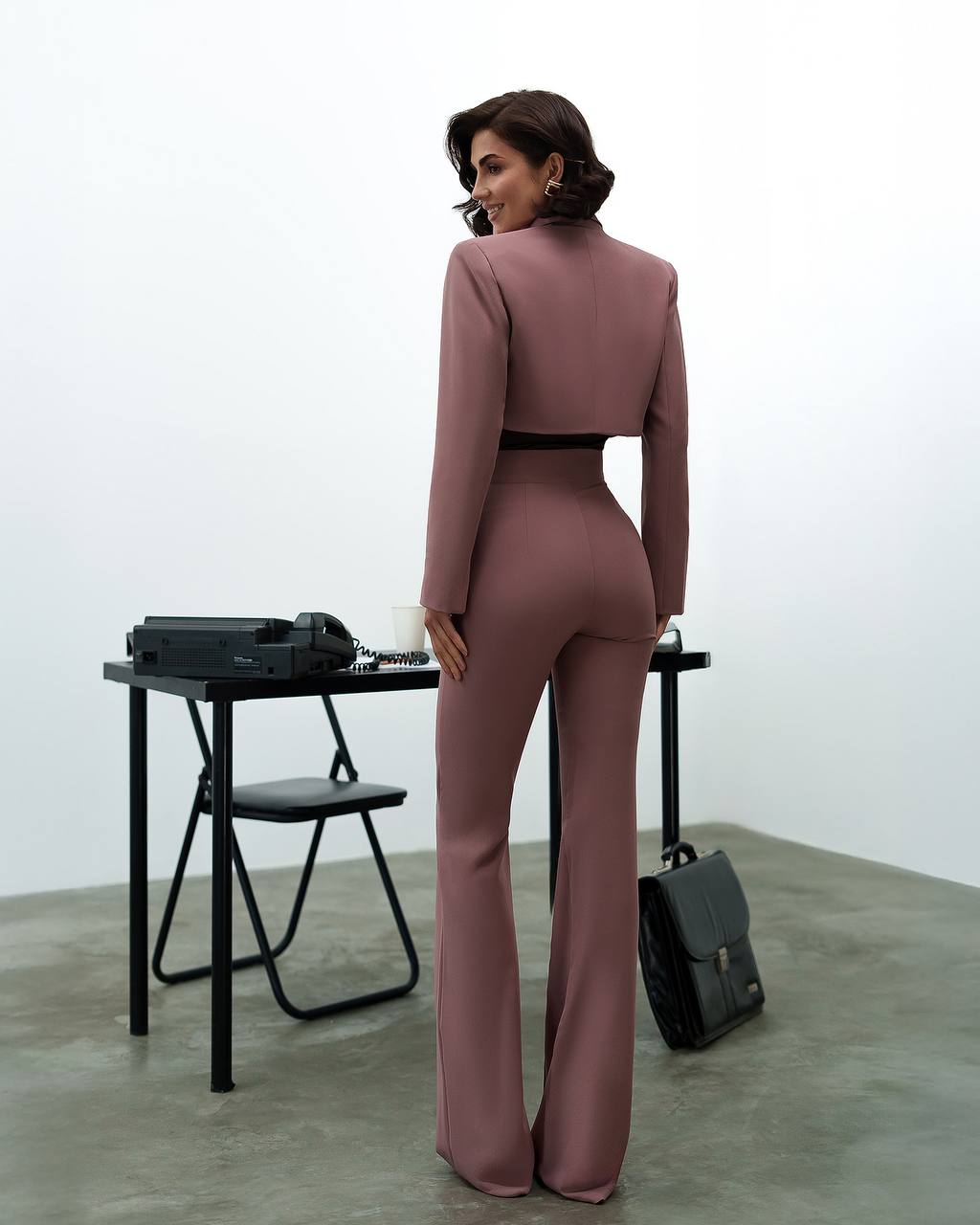 Woman in a mauve jumpsuit standing in a minimalistic room with a table and chair.