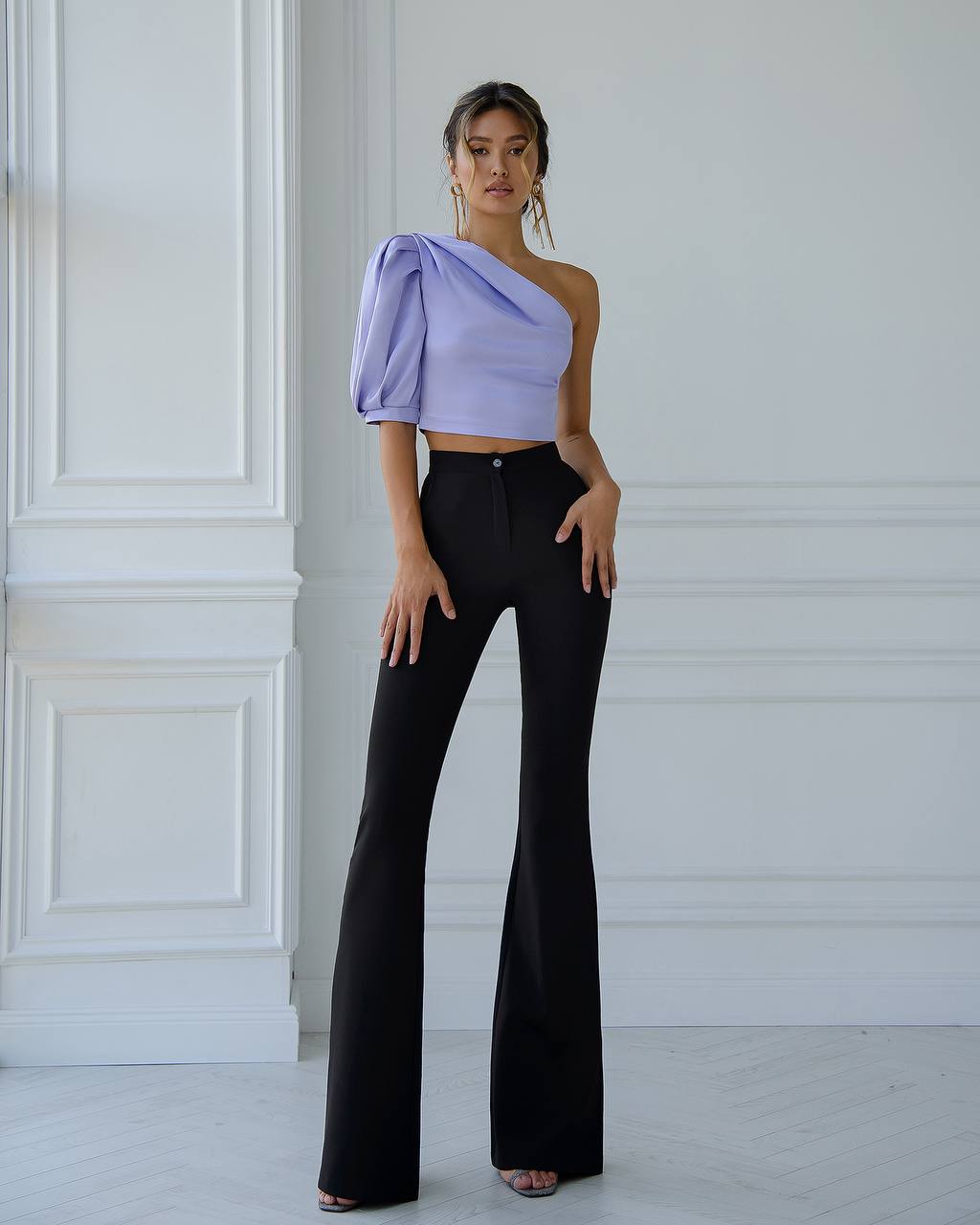 Woman wearing a one-shoulder lavender top and black flared pants in a white room.