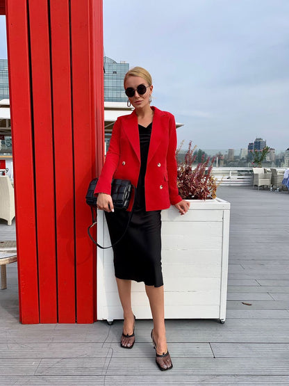 Woman in a red blazer and black skirt standing next to a red wall with a cityscape in the background.