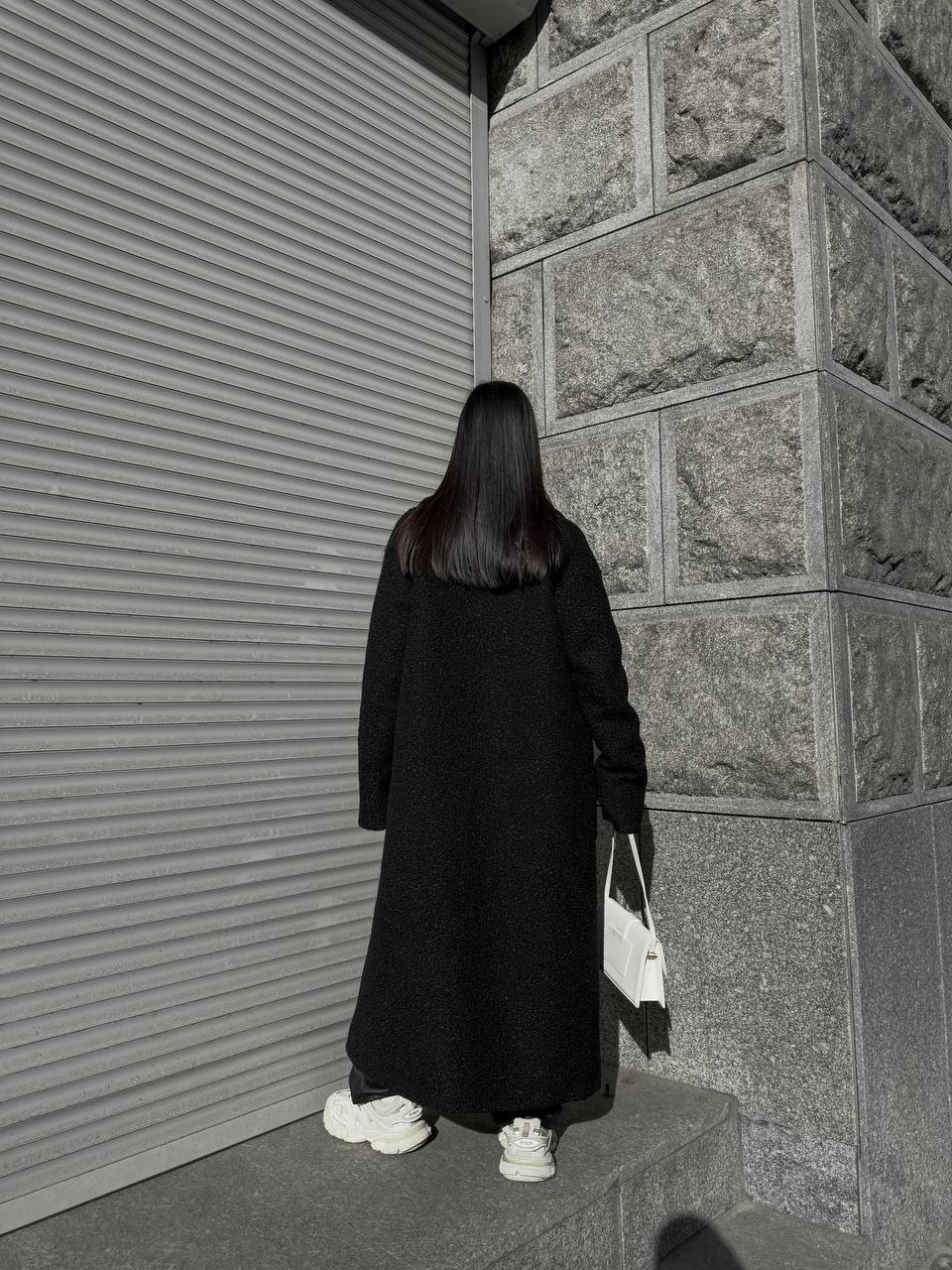 Person in a long black coat standing on steps against a textured wall.