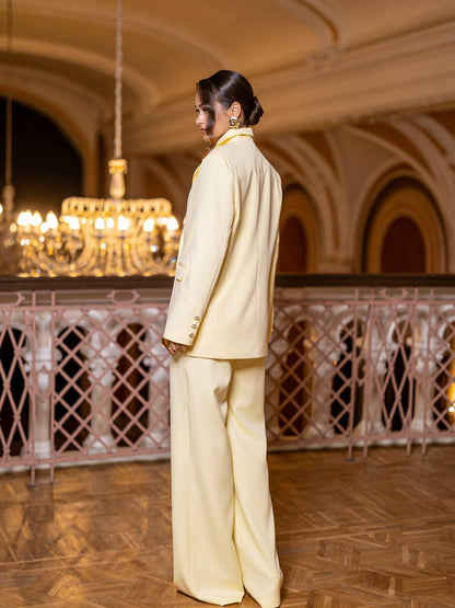 Person wearing a light-colored suit standing in an elegant room with a chandelier.
