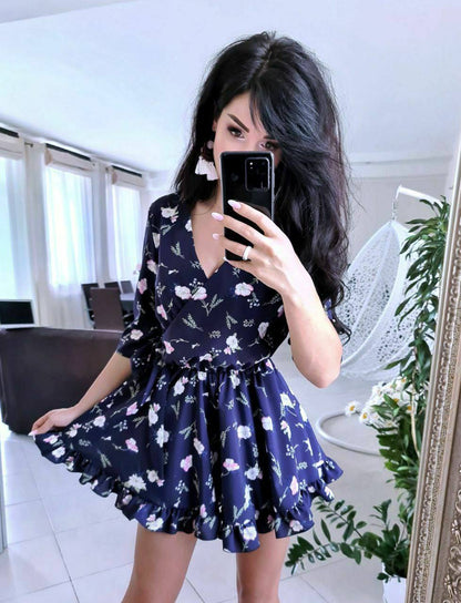 Woman taking a mirror selfie wearing a navy floral dress in a room with white walls and a plant.