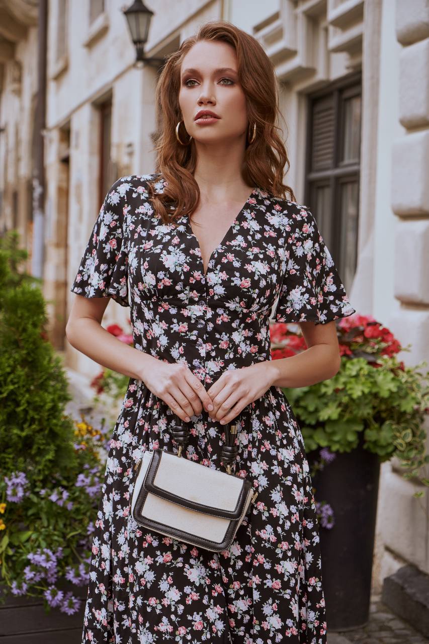 Woman in a floral dress holding a white handbag on a street with plants and buildings in the background